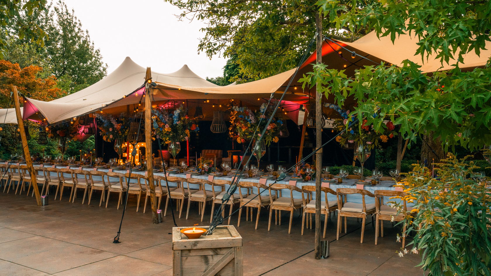 Een elegante en feestelijk gedekte dinertafel voor een 30ste verjaardag bij restaurant De Luytervelde, georganiseerd door een high-end wedding en event planner. De lange tafel staat onder een luxe tent, rijkelijk versierd met kleurrijke bloemen en sfeervolle verlichting, omgeven door weelderig groen voor een intieme en verfijnde ambiance.
